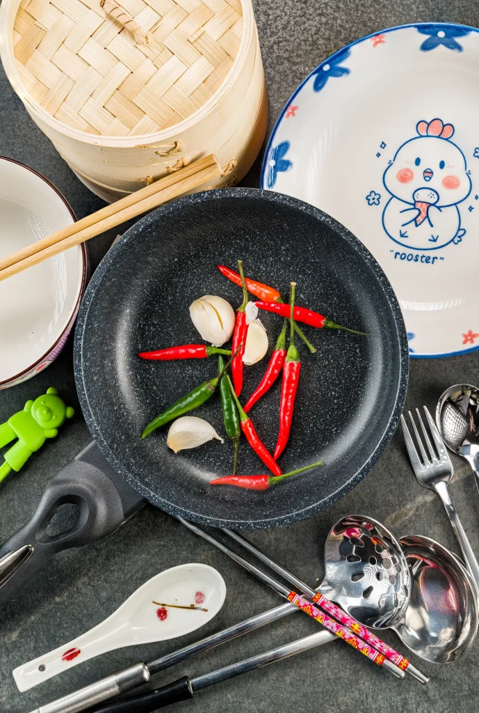 A pan with chili peppers and garlic cloves on a kitchen counter surrounded by various cooking utensils and a bamboo steamer.