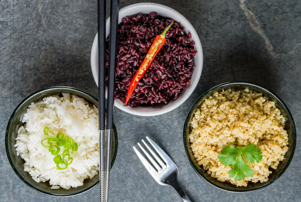 Three bowls of different grains: white rice garnished with green onions, black rice with a chili pepper, and quinoa garnished with herbs, accompanied by a pair of chopsticks and a fork.