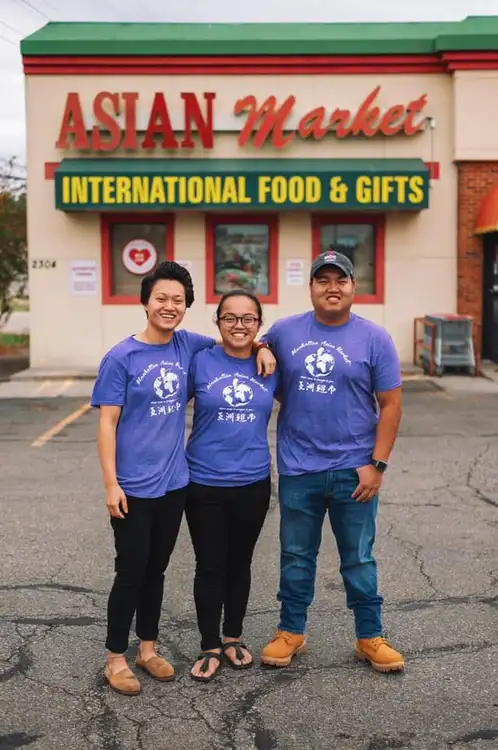 Three smiling individuals standing in front of the "asian market international food & gifts" store, wearing matching blue t-shirts with logos, possibly indicating they are staff or part of a team event.