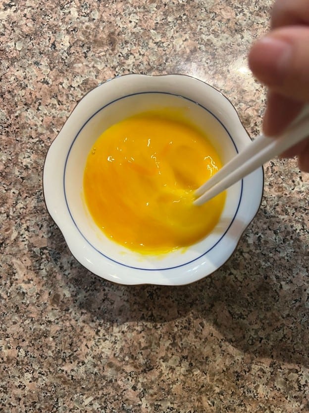 A person's hand whisking egg yolks in a white and blue patterned bowl on a granite countertop, as seen on a website.