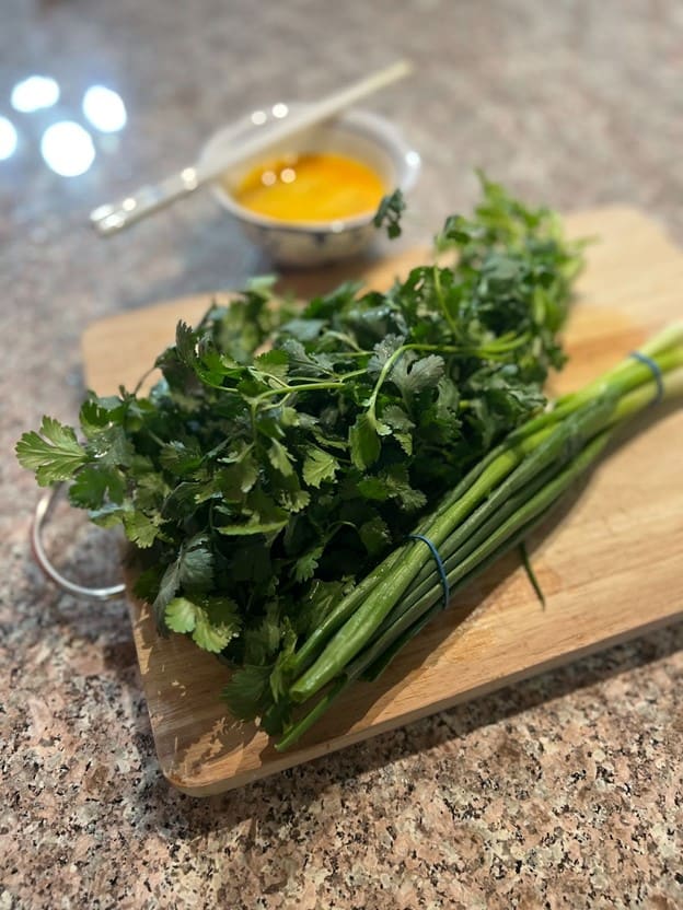 Fresh cilantro and green onions arranged on a wooden cutting board with a bowl of sauce in the background, ready for cooking preparation, presented by MamaFangs.com.