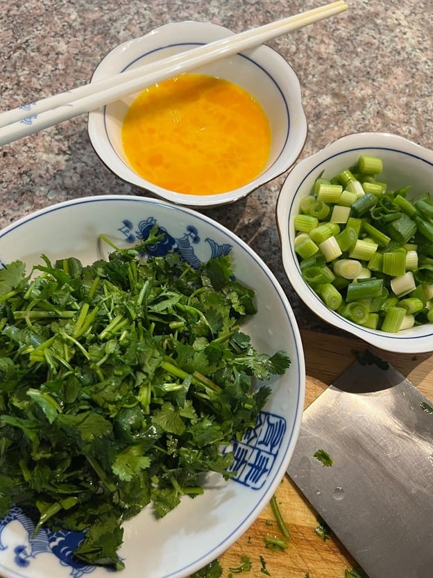 A bowl of chopped fresh cilantro, a bowl of sliced green onions, and a bowl of beaten egg, with a pair of chopsticks resting on a spoon, on a kitchen counter ready for cooking