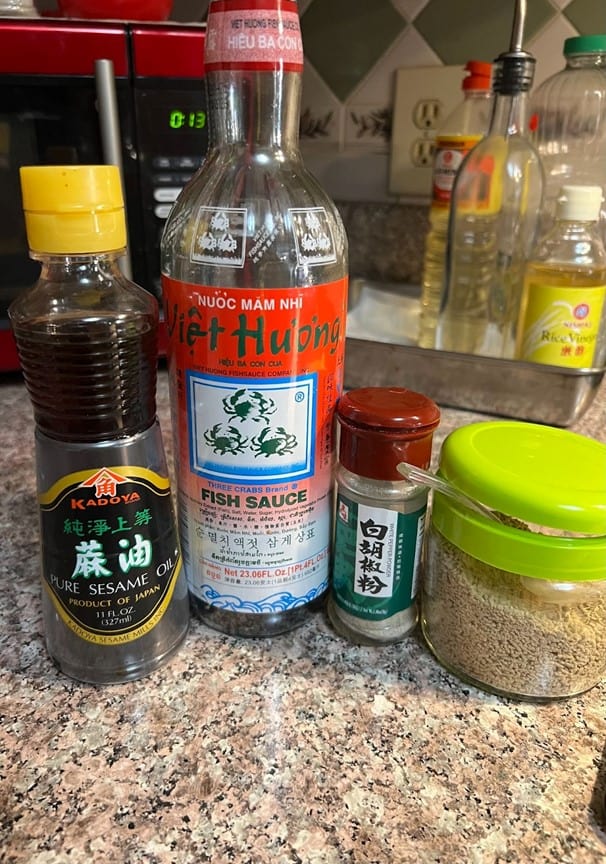 A collection of Asian condiments and spices from MamaFangs.com on a kitchen countertop, featuring fish sauce, sesame oil, a small jar of spice, and a container of what appears to be