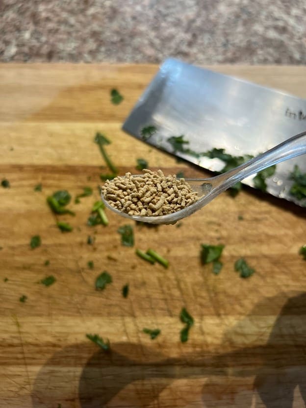 A teaspoon of sesame seeds held above a wooden cutting board sprinkled with chopped herbs and a chef's knife in the background, captured by MamaFangs.com.