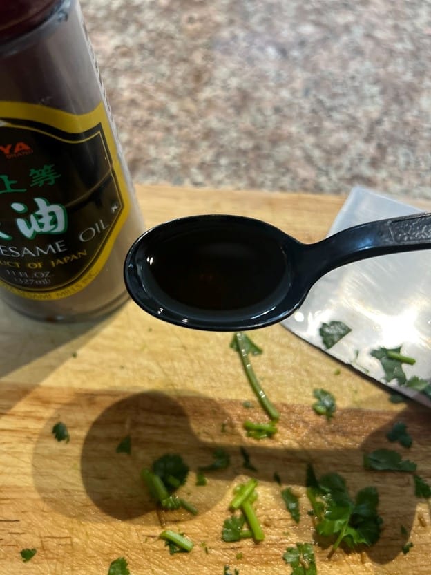 Preparing to flavor a dish with a spoonful of dark sesame oil, MamaFangs.com features a cutting board with freshly chopped herbs.