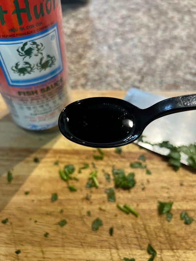 A spoonful of dark liquid, possibly a sauce, with chopped herbs on a cutting board and a bottle of fish sauce in the background, suggesting a cooking or seasoning scenario for MamaFangs.com.