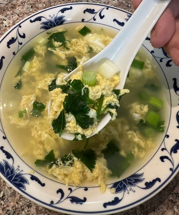 A bowl of homemade egg drop soup from MamaFangs, garnished with green onions and cilantro, served with a spoonful ready to enjoy.