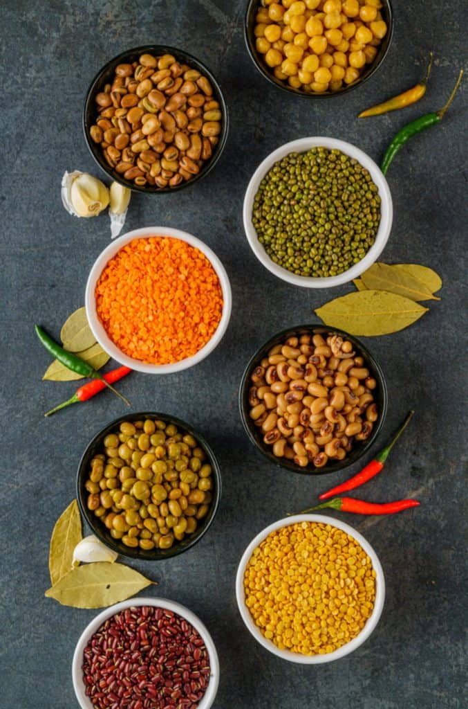 Assorted legumes and spices arranged in bowls on a dark textured surface, featuring a colorful variety of beans, lentils, chickpeas, and vibrant chili peppers, with bay leaves and garlic cloves