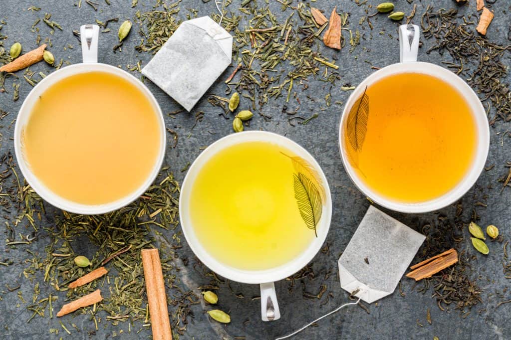 Three cups of tea with different hues ranging from pale yellow to golden, surrounded by an assortment of loose tea leaves and spices on a textured grey surface. Each cup has a tea bag inside or beside it