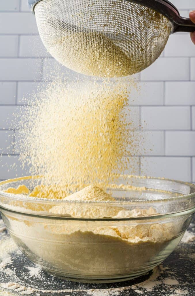 A person sifting flour through a fine mesh sieve into a glass mixing bowl, with a light dusting of baking ingredients on the work surface below.