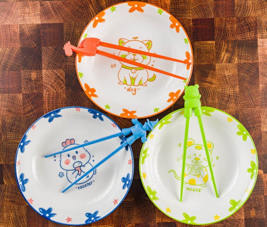 Set of colorful children's plates with animal illustrations, accompanied by matching utensils with playful tops, arranged on a patterned tablecloth, perfect for enhancing your household items collection.