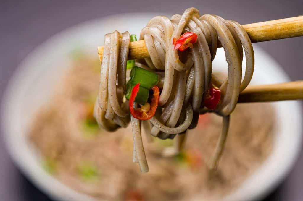 Cooking soba noodles garnished with slices of chili and green leaves, tantalizingly suspended between chopsticks with a blurred bowl in the background.