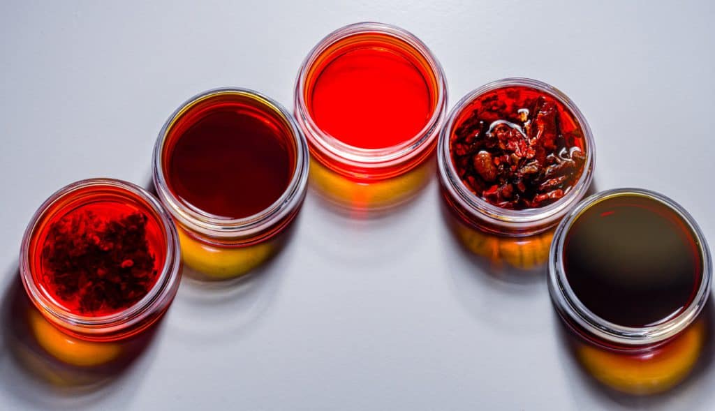Five glass jars filled with various shades of red and amber cooking sauces casting colorful reflections on a white surface.