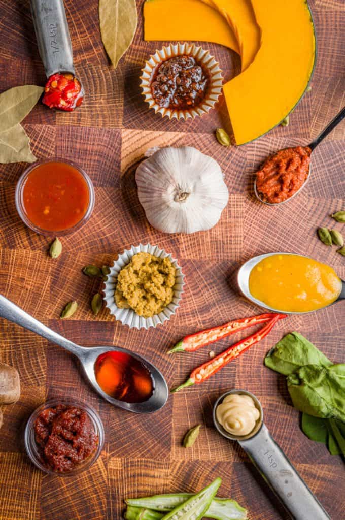 A vibrant array of spices and ingredients artfully arranged on a wooden surface, featuring sliced pumpkin, garlic, red chilies, and various cooking sauces in spoons and small dishes, ready for an adventurous