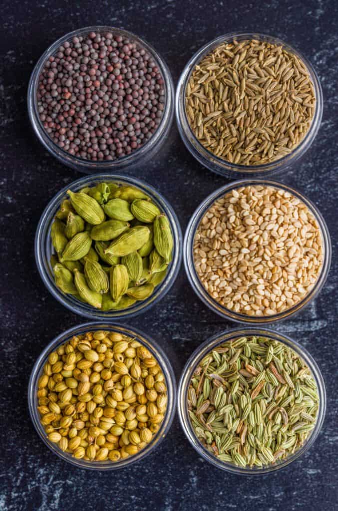 Assorted gourmet spices and seeds in small glass jars arranged on a dark textured surface.
