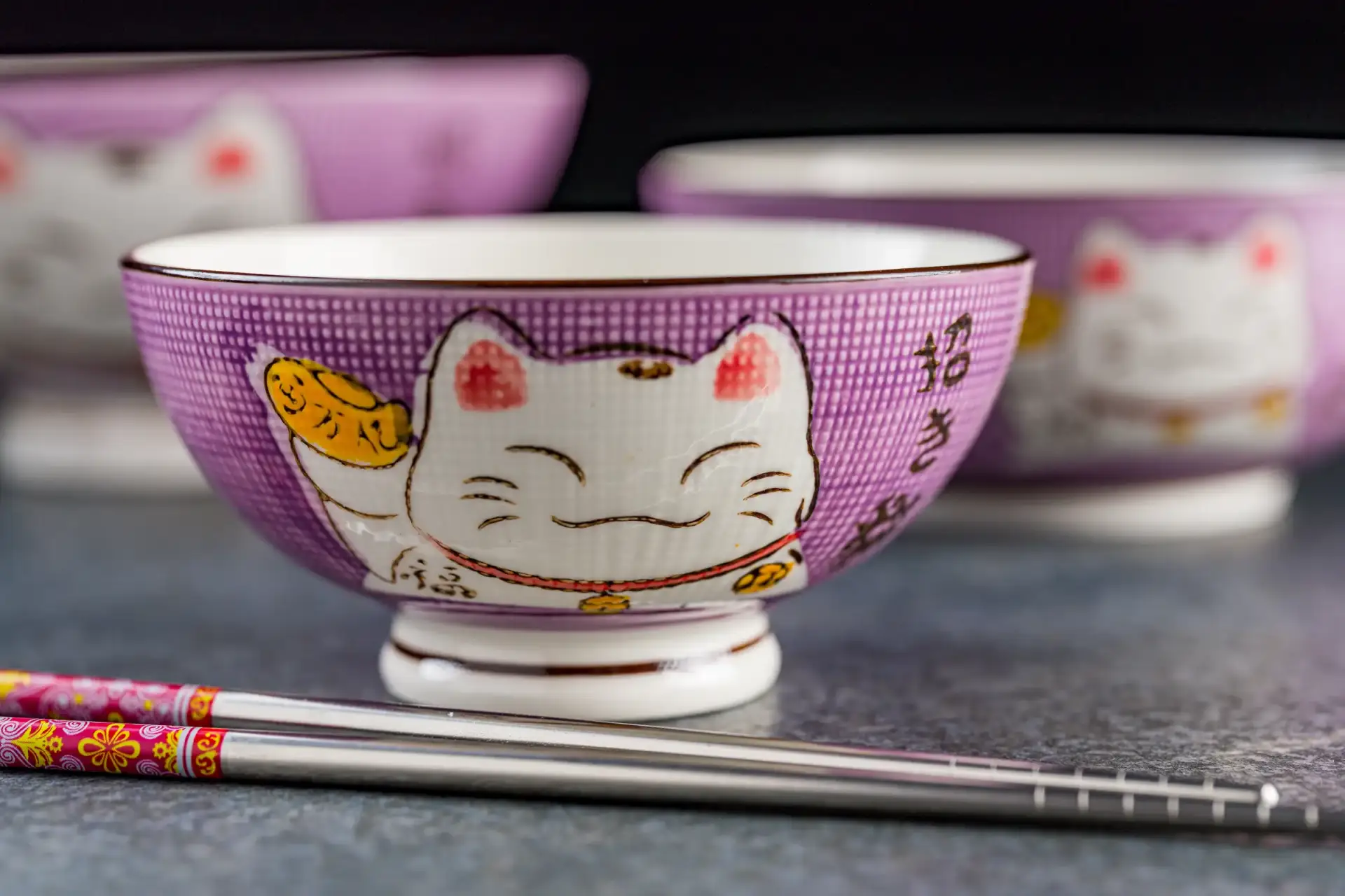 Close-up of a decorated cat-themed rice bowl with chopsticks in front, other bowls blurred in the background.