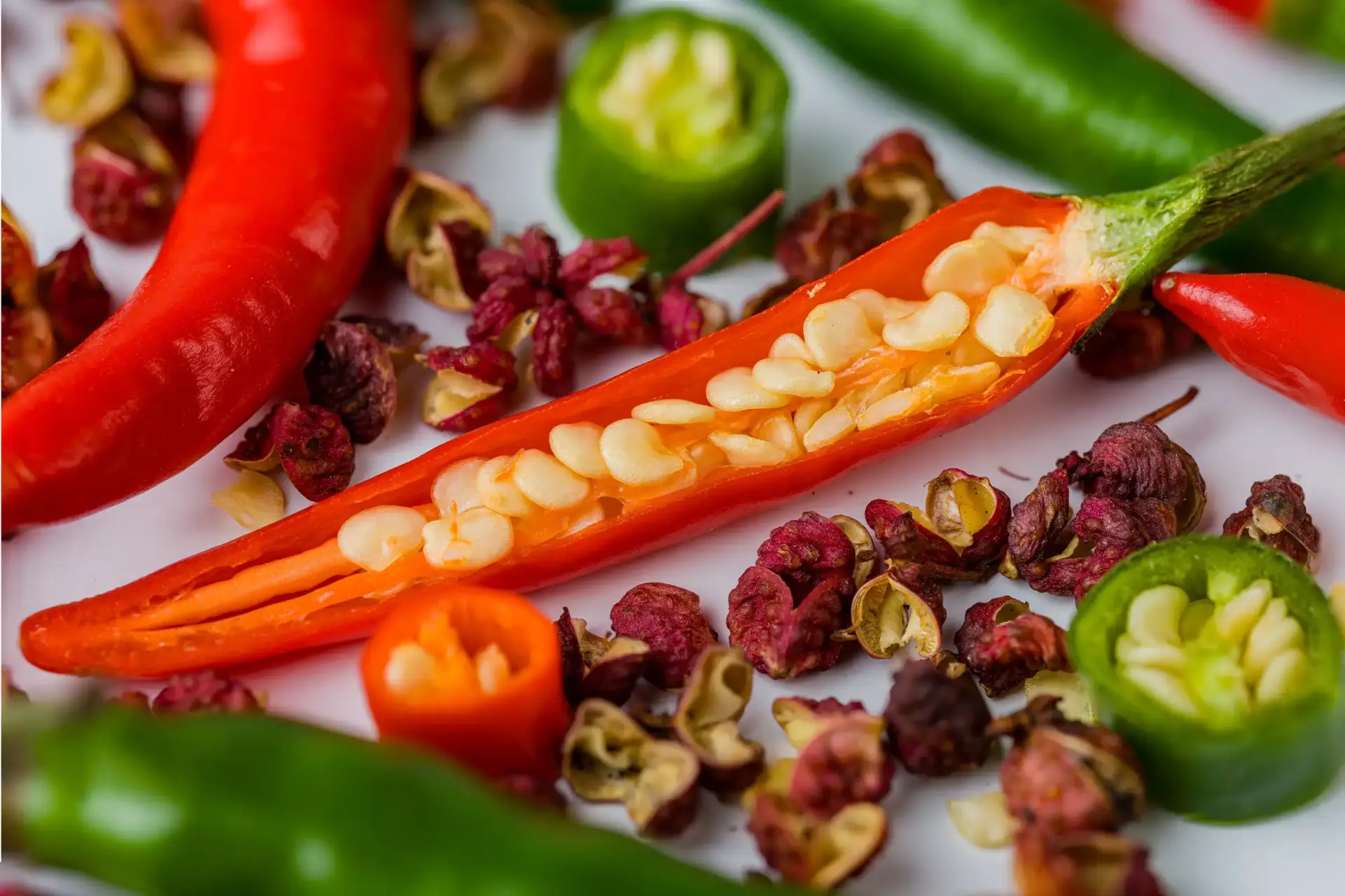 Sliced and whole chili peppers with visible seeds on a white surface, surrounded by dried pepper flakes.