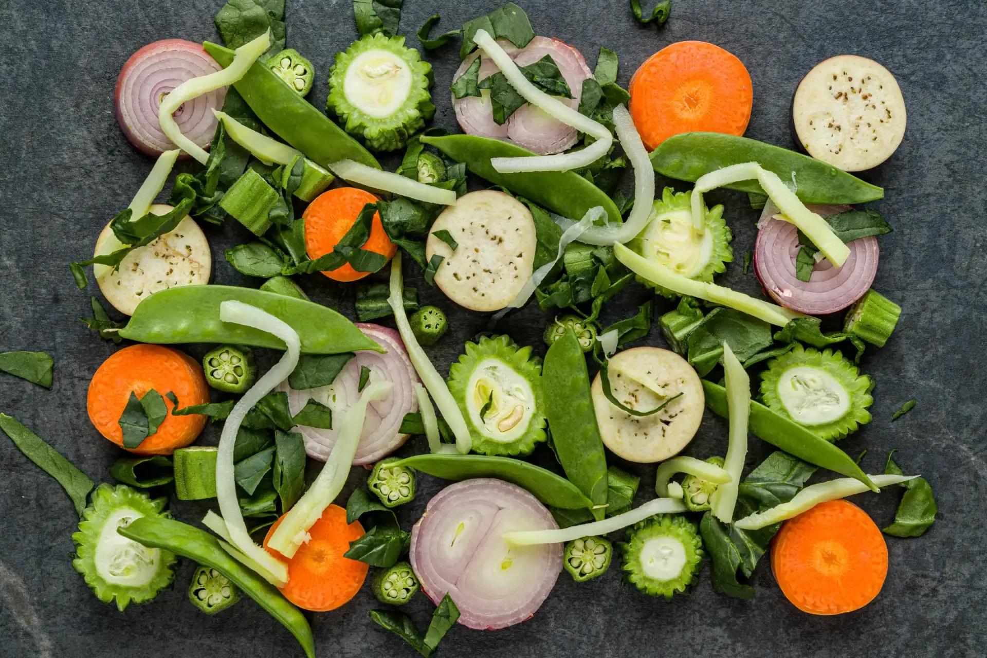 Assorted sliced vegetables arranged on a dark slate surface.